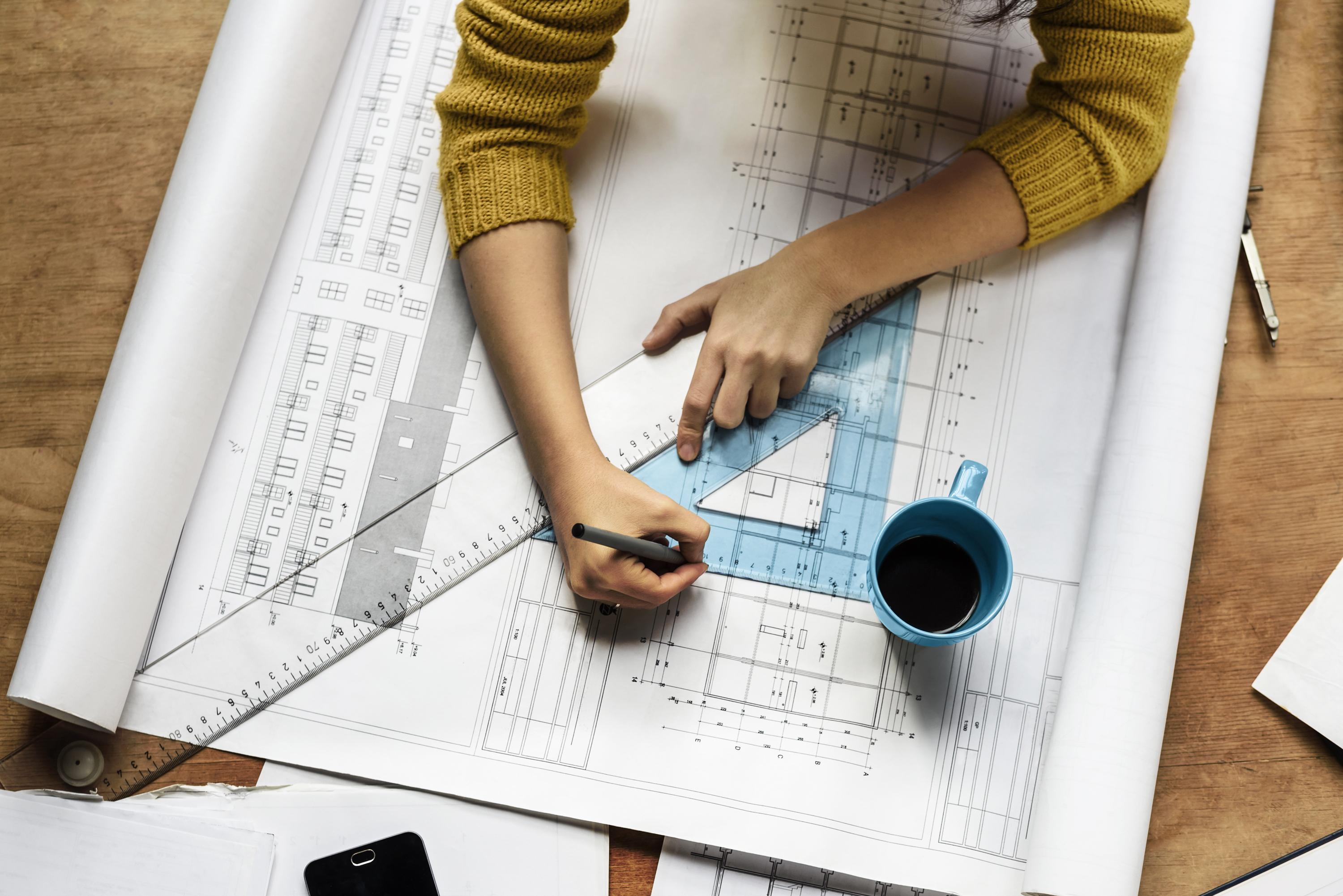 A close-up of a person's hands working on architectural blueprints, using an arc triange and pen, with a cup of coffee nearby.