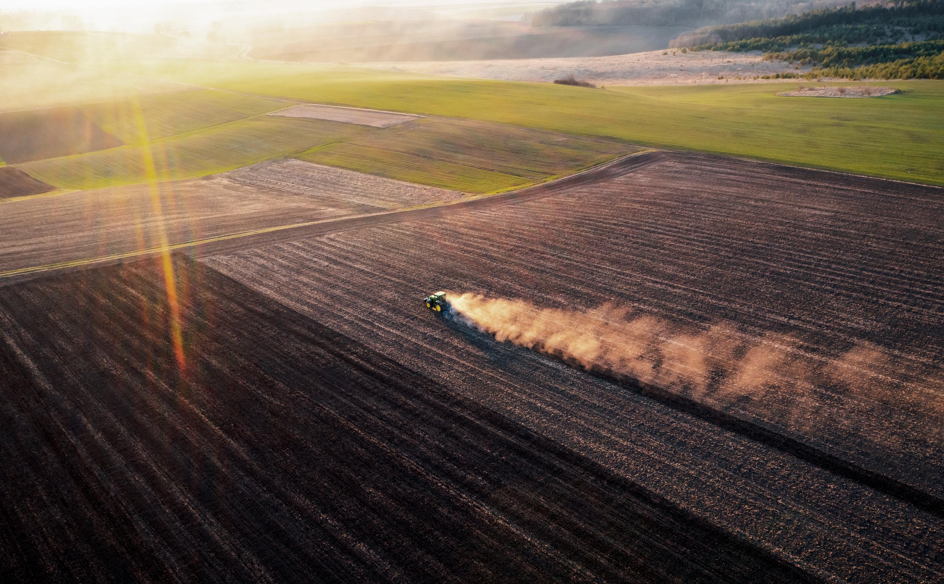 A wide-angle aerial shot of a tractor plowing through a field during sunset, leaving a trail of dust in a farm landscape.