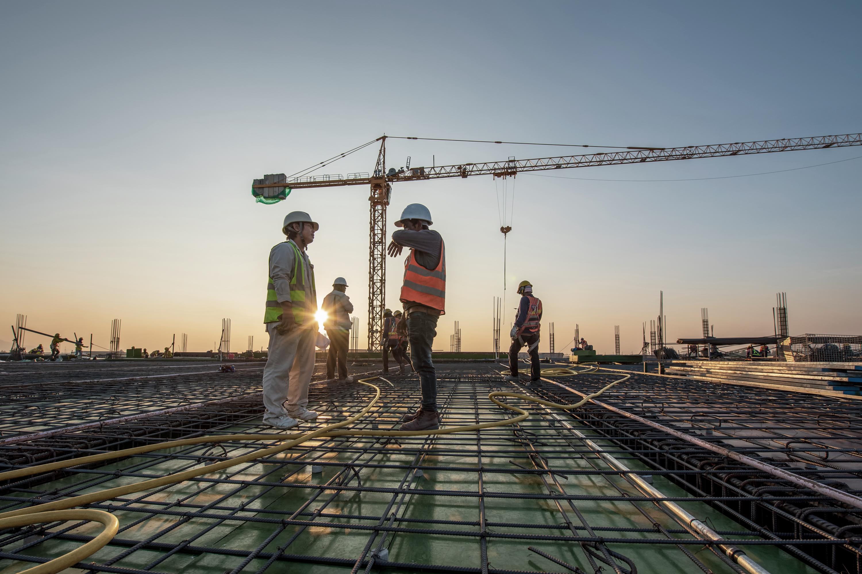 Construction workers in helmets and safety gear standing on a building site with cranes and scaffolding in the background. 