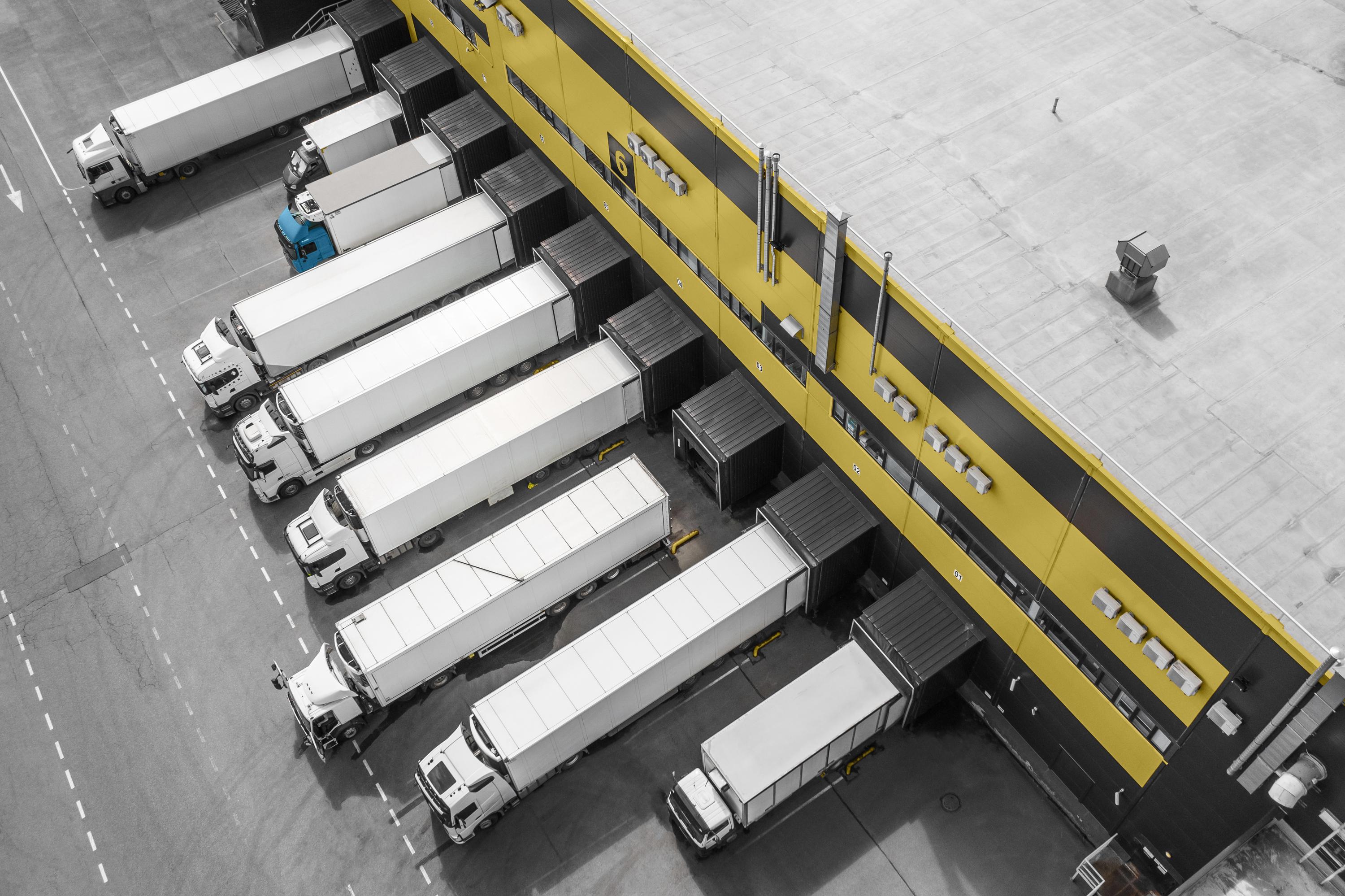 Overhead view of a large industrial facility with rows of parked white cargo trucks and containers. 